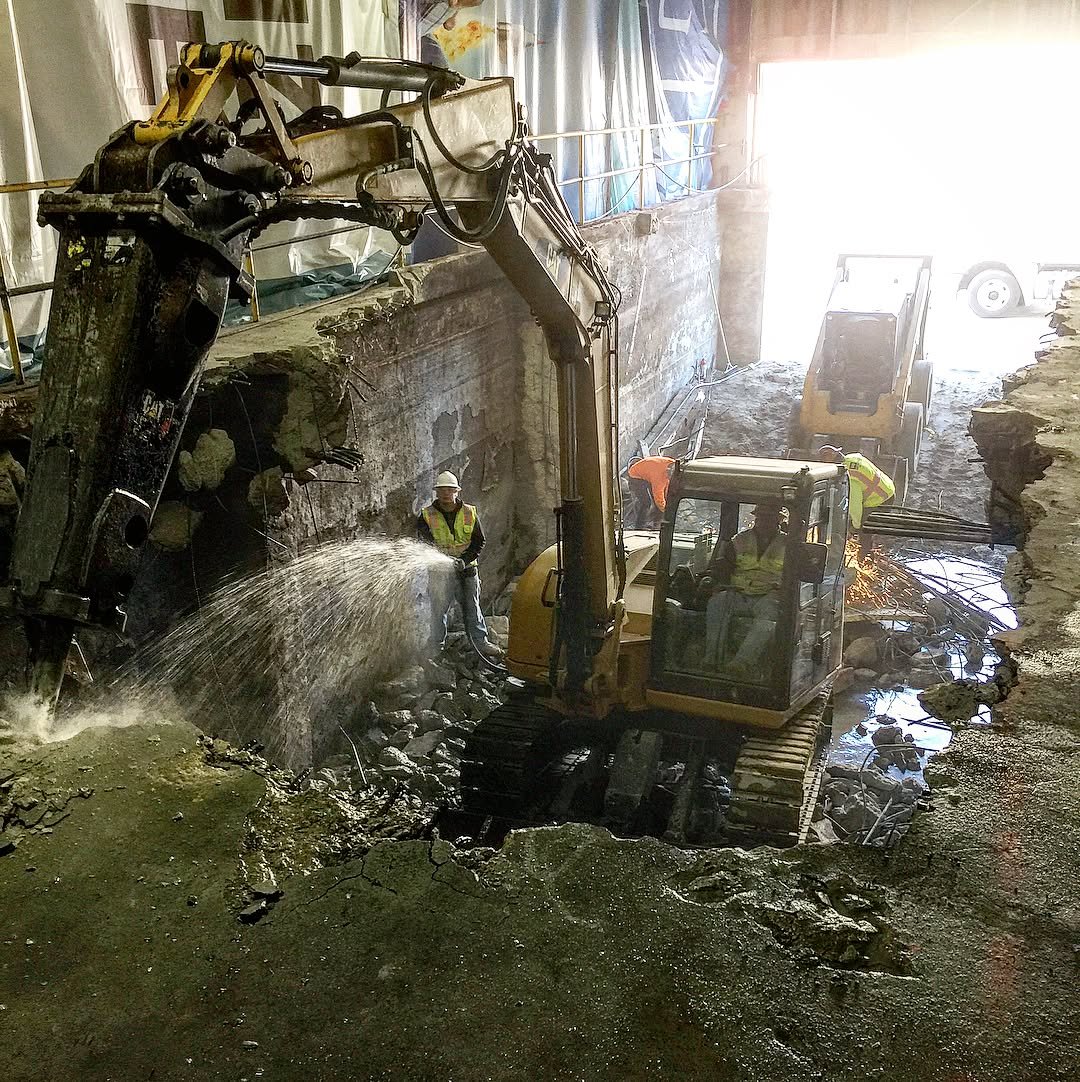 Mini excavator with hoe ram breaking concrete in dramatic interior — crew member visible with water suppression active