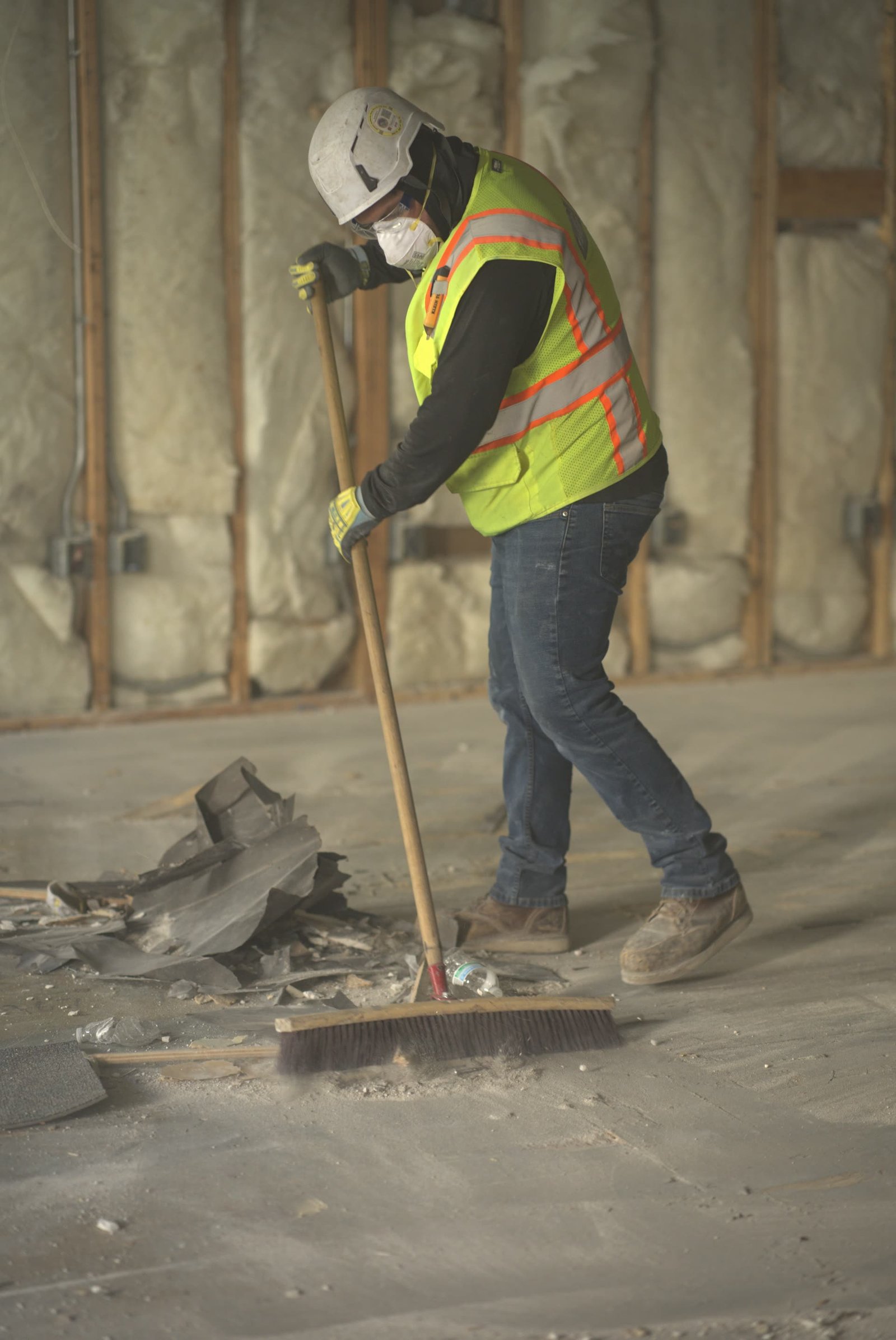 DSI laborer in full PPE sweeping demolition debris on concrete floor of stripped commercial building