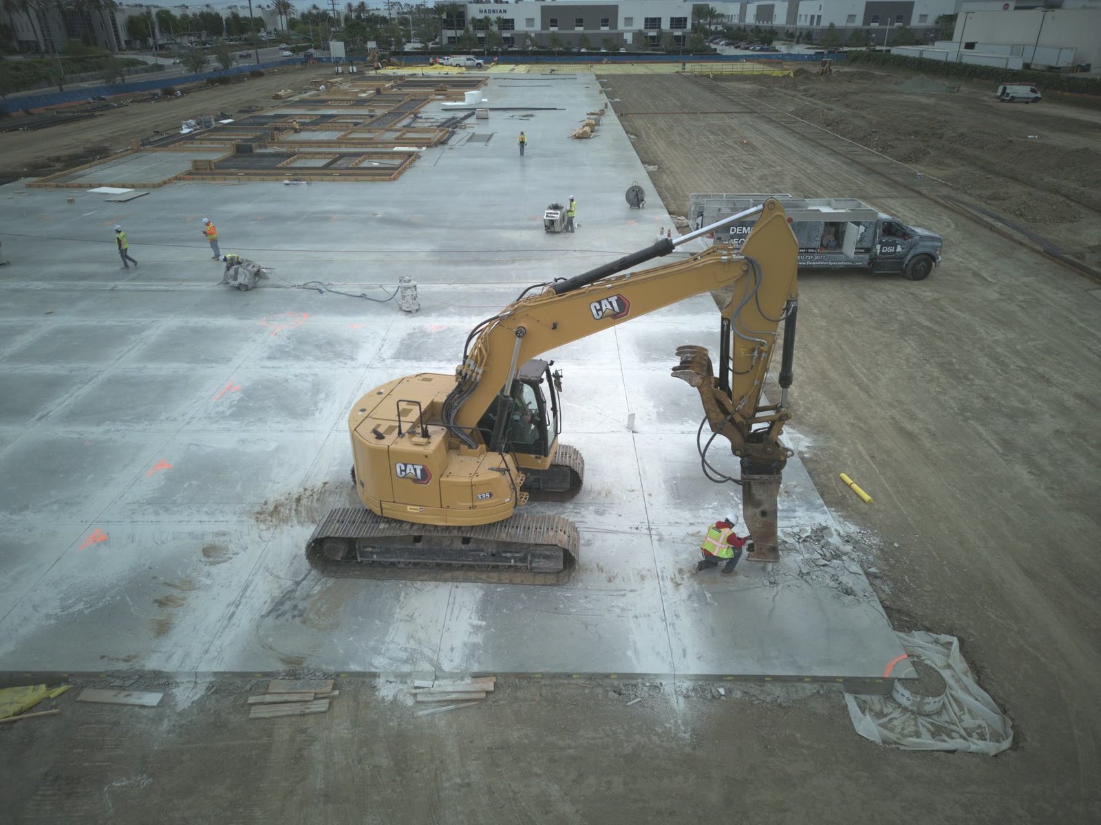 Aerial drone view of DSI CAT excavator with hoe ram breaking large commercial concrete slab with DSI truck on site